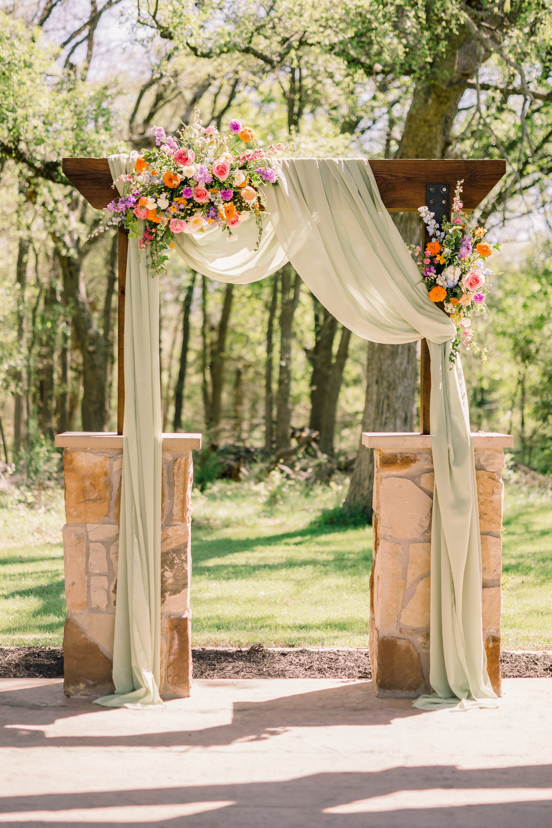 Draped wooden arch at The Springs Alvarado with sage fabric and bright spring florals of coral, yellow, and blue on stone pedestals, mature trees behind, by B.blumen