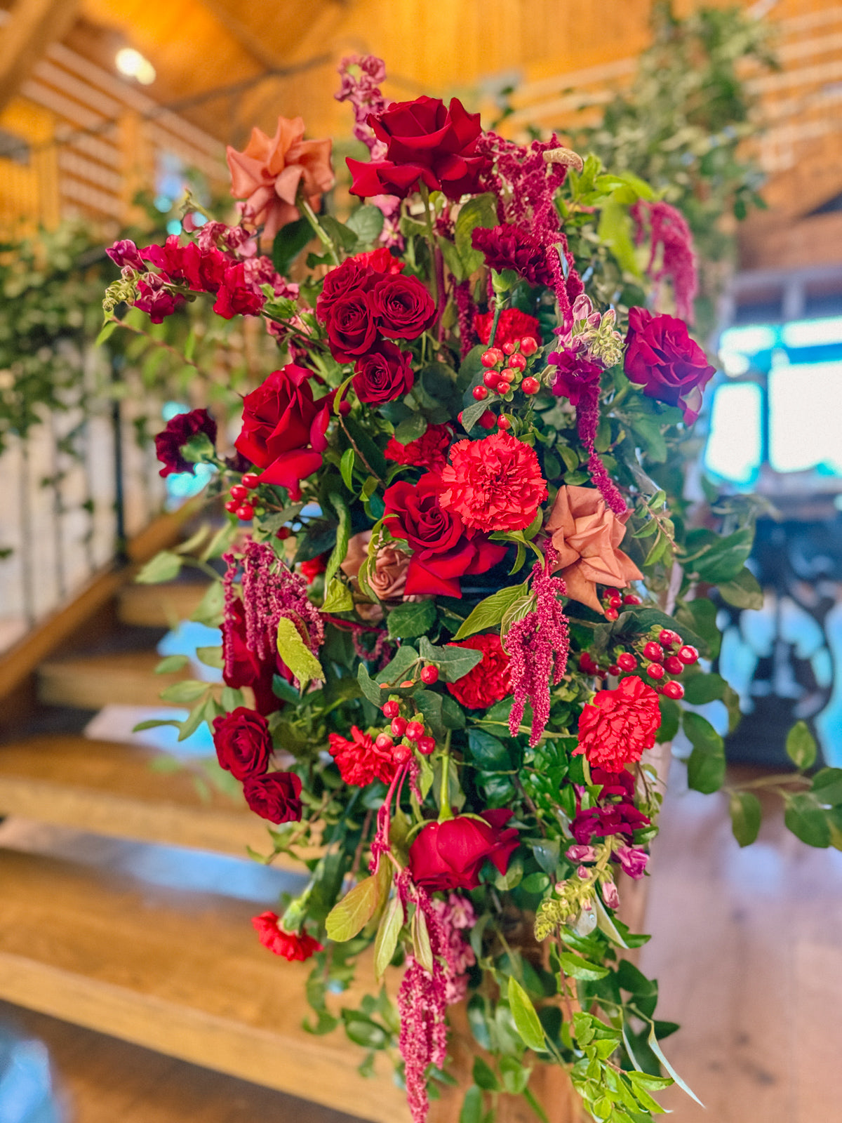 Tall arrangement of deep red roses, burgundy carnations, blush garden roses, and hanging amaranthus at the base of the staircase at Timberview Lodge, by B.blumen