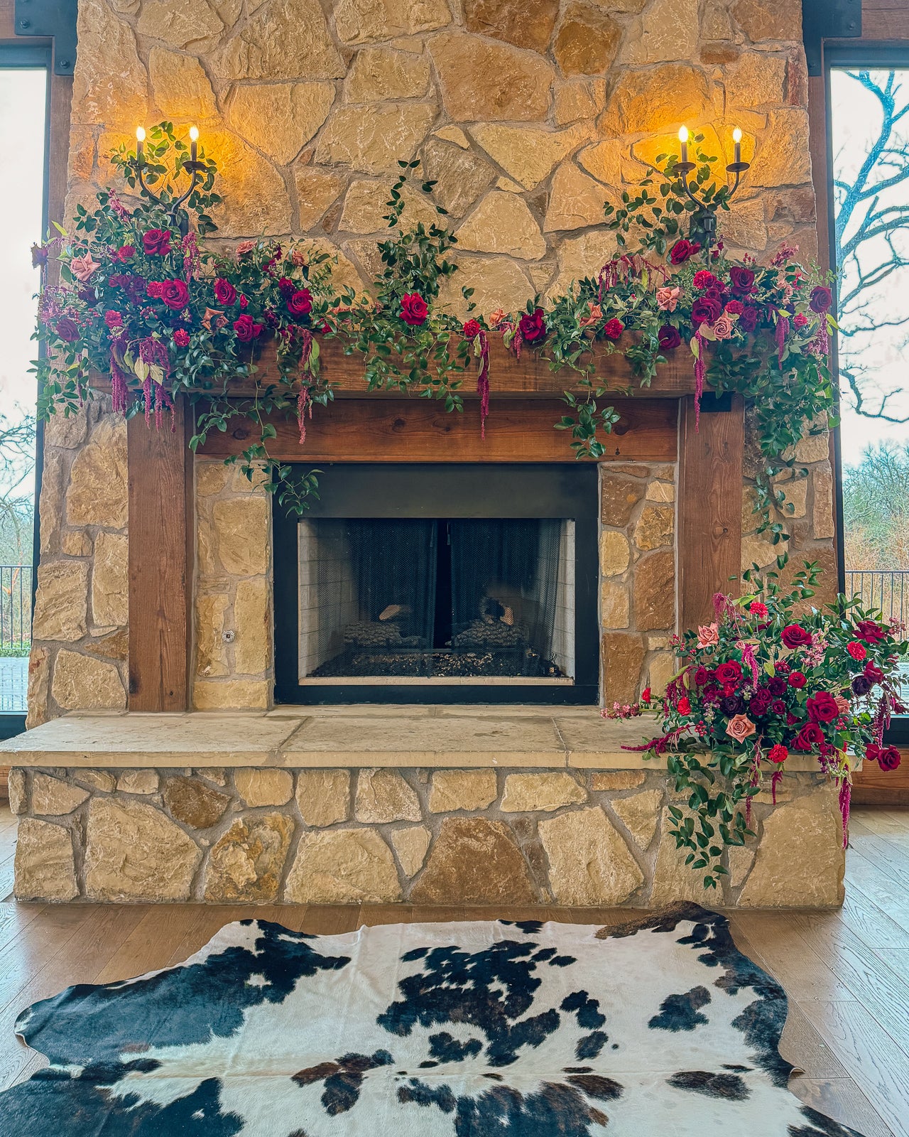 Stone fireplace at Timberview Lodge dressed with a burgundy mantel garland of deep red roses, carnations, and trailing greenery, paired with a cascading side arrangement and a cowhide rug on the wood floor, by B.blumen