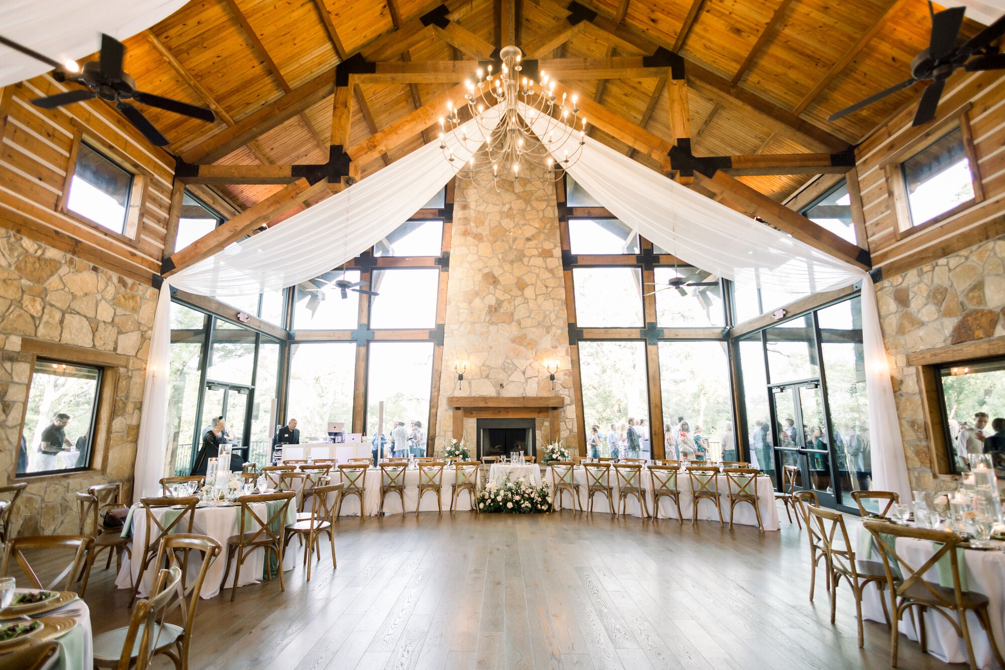 Interior of Timberview Lodge at The Springs Alvarado, cathedral ceiling with iron chandelier, three-story stone fireplace, draped fabric, and the head table set with low white centerpieces, by B.blumen