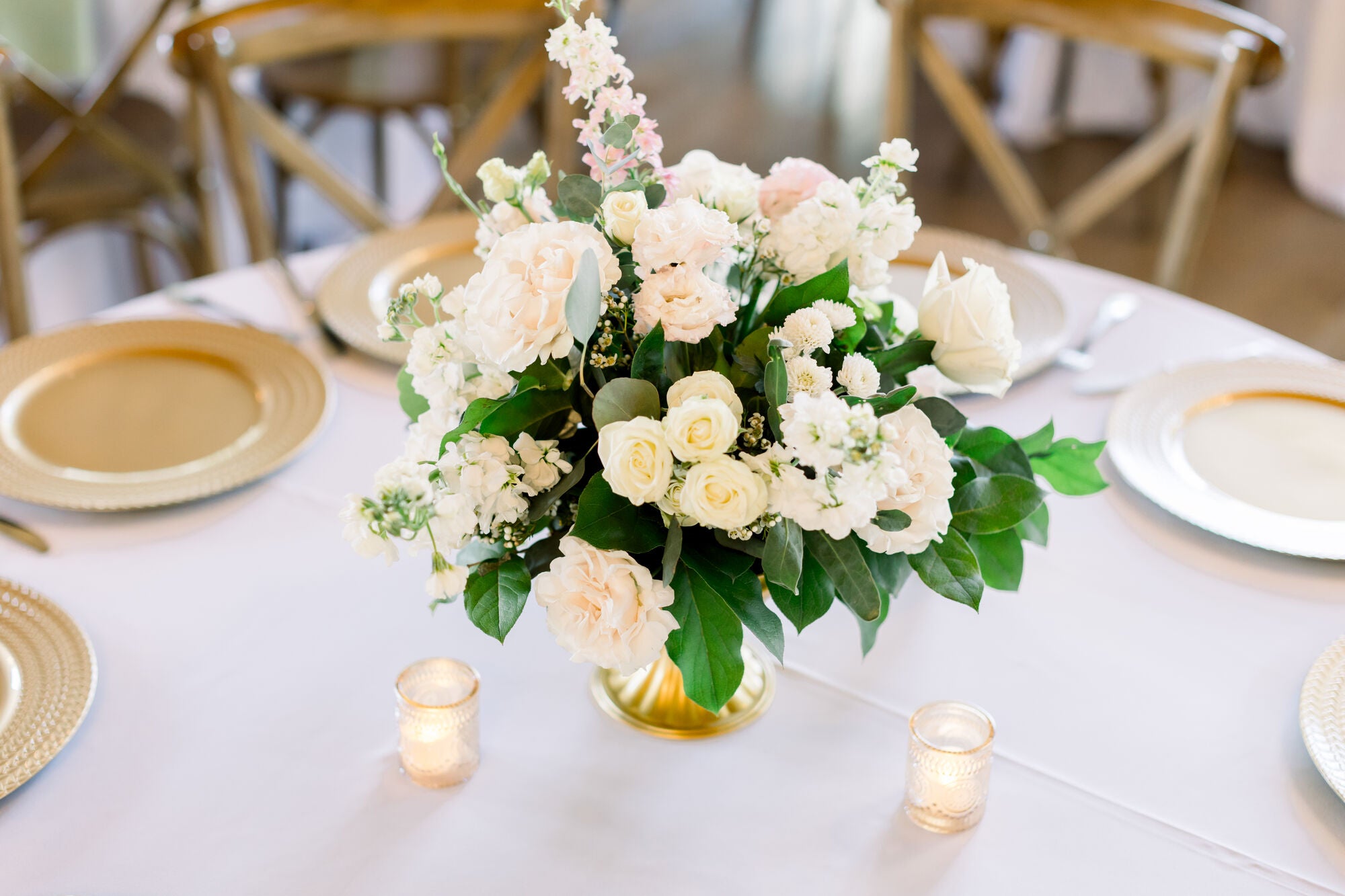 Round reception table at Timberview Lodge with a low white-and-blush centerpiece, gold chargers, and wood crossback chairs, by B.blumen