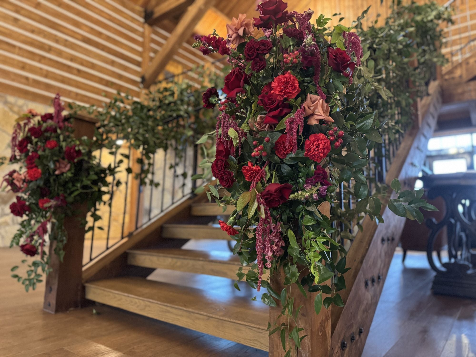 Sweeping floor arrangements of deep red roses, burgundy carnations, blush garden roses, and hanging amaranthus flanking the wood-and-iron staircase at Timberview Lodge, by B.blumen