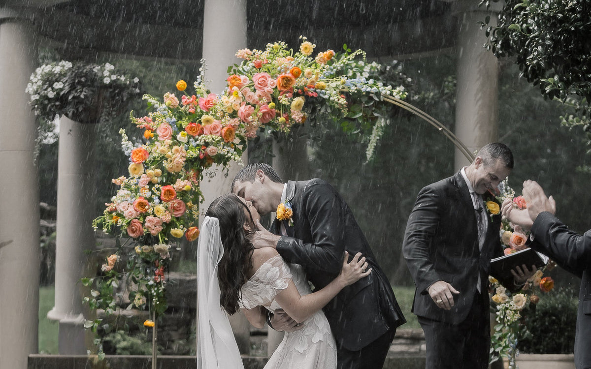 Bride and groom kiss under a floral arch in the rain.