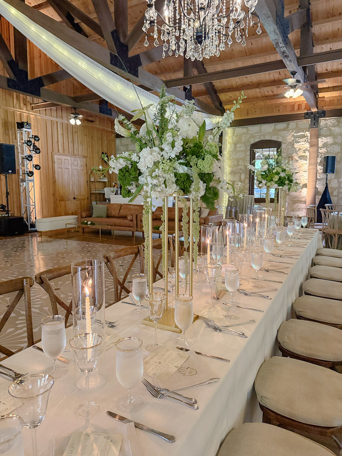 Tall pedestal centerpiece rising from a brass candelabra stand on the long head table at Poetry Springs, white roses, ranunculus, viburnum, and hanging amaranthus, gold cylinder votives along the runner, wood-beam pavilion overhead, by B.blumen