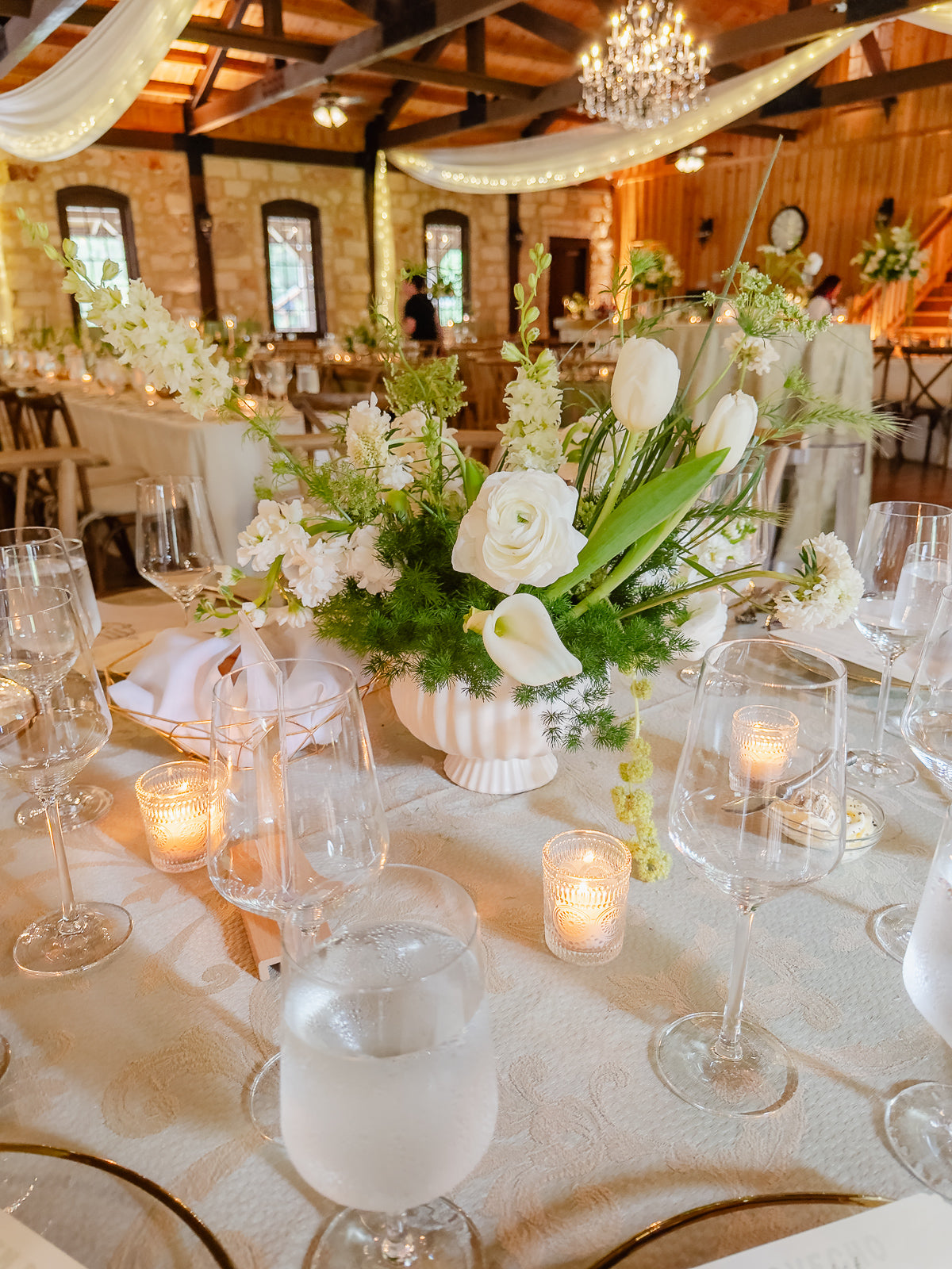 Round table close-up at Poetry Springs with a white fluted compote vase holding tulips, callas, ranunculus, and asparagus fern, votives glowing, brocade-pattern linen, by B.blumen