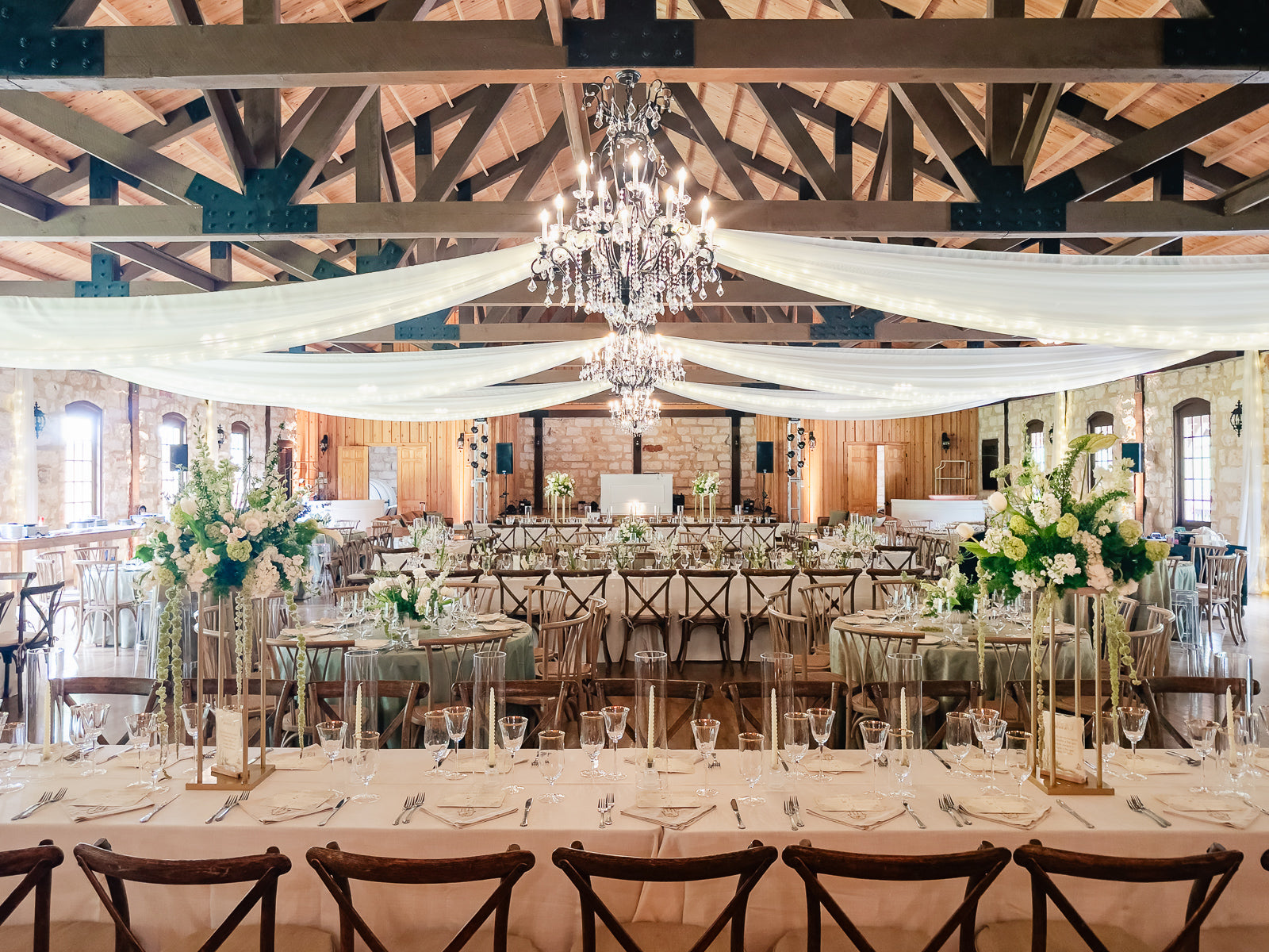 Wide reception establishing at Poetry Springs with the dark exposed truss ceiling draped in white fabric and twinkle lights, crystal chandelier center, head table in the distance, round tables with tall pedestal florals in the foreground, by B.blumen