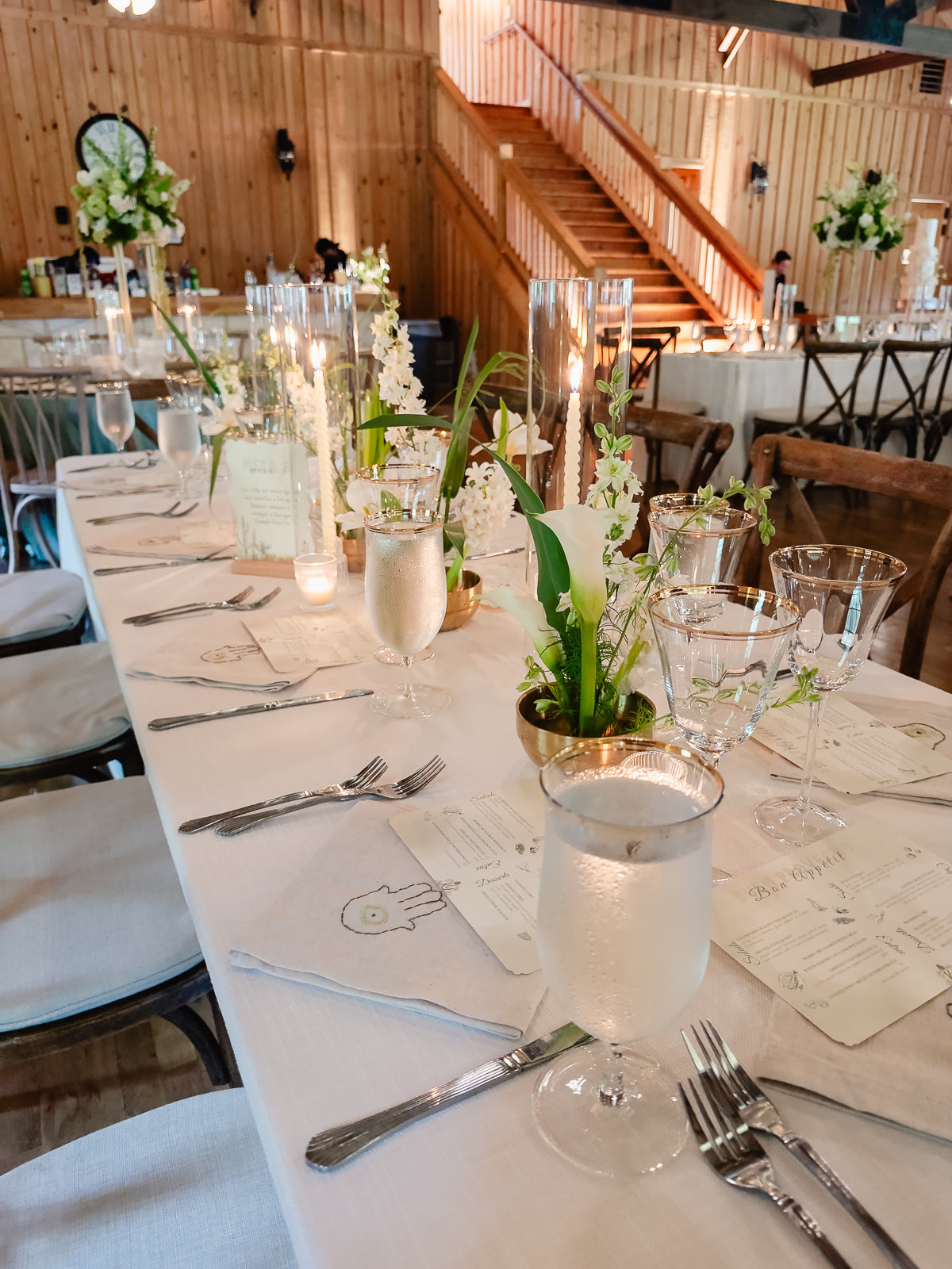 Long head table at Poetry Springs with gold cylinder vases holding single calla lily stems, white tapers in candleholders, gold-rimmed goblets, calligraphy menu cards, wood staircase in the background, by B.blumen