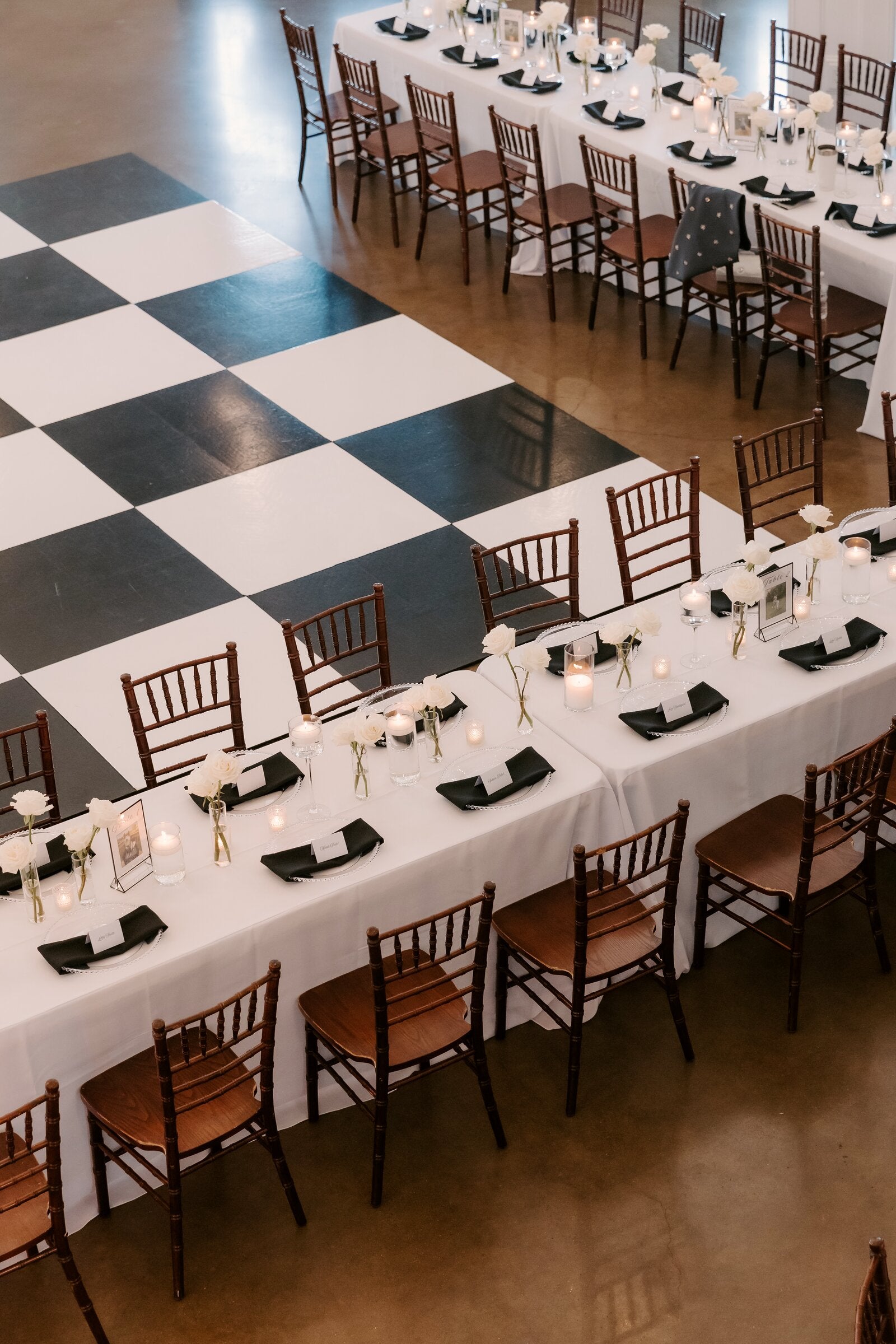 Long reception table at Rockwall Manor running along the edge of the checkerboard dance floor with low white floral centerpieces, black napkins, and chiavari chairs, by B.blumen