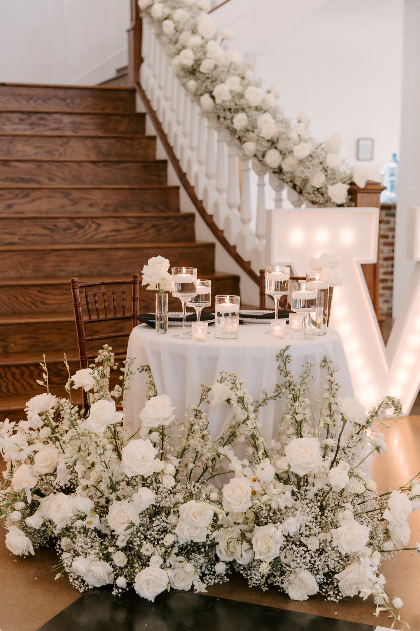 Sweetheart table at the base of the Rockwall Manor staircase with a flowing baby's breath garland down the banister, a marquee letter at the side, and a lush white ground arrangement at the table's base, by B.blumen