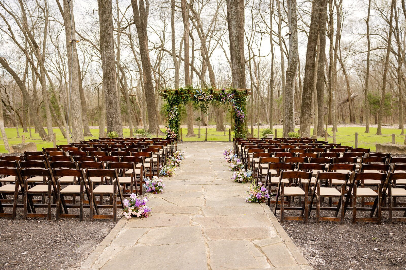 Outdoor ceremony space at The Springs Waxahachie with rows of wooden chairs facing an ivy-covered pergola altar by B.blumen