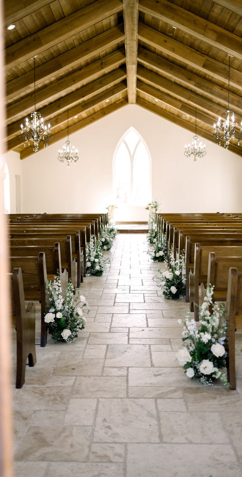 Interior of The Springs Waxahachie chapel with wooden pew aisle, gothic-arched window, and white floral aisle markers by B.blumen