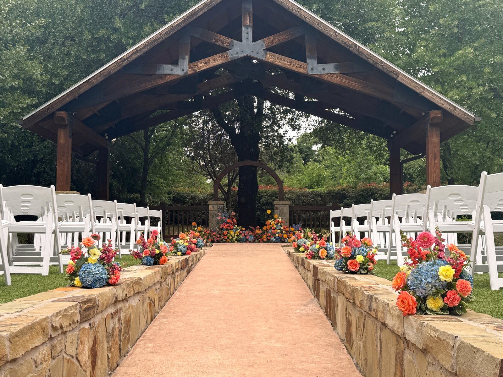 Wood pavilion outdoor ceremony at The Springs Weatherford Westwood Hall, with stone aisle walls and vibrant orange, blue, and yellow florals by B.blumen