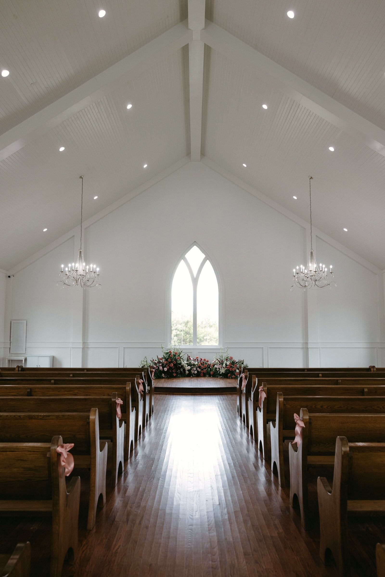 Interior of The Springs Weatherford chapel with vaulted ceiling, gothic arched window, chandeliers, pink pew ribbons, and a sweeping altar arc of dusty pink and burgundy florals by B.blumen