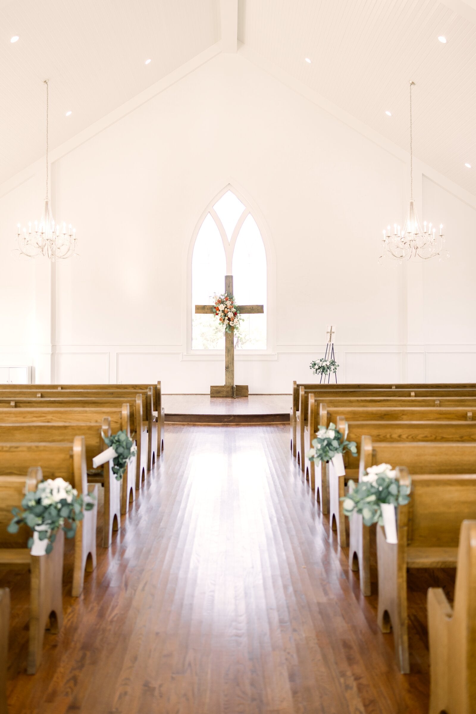 Chapel altar at The Springs Weatherford with wood cross and floral arrangement framed by the gothic arched window, eucalyptus aisle markers down the pews by B.blumen