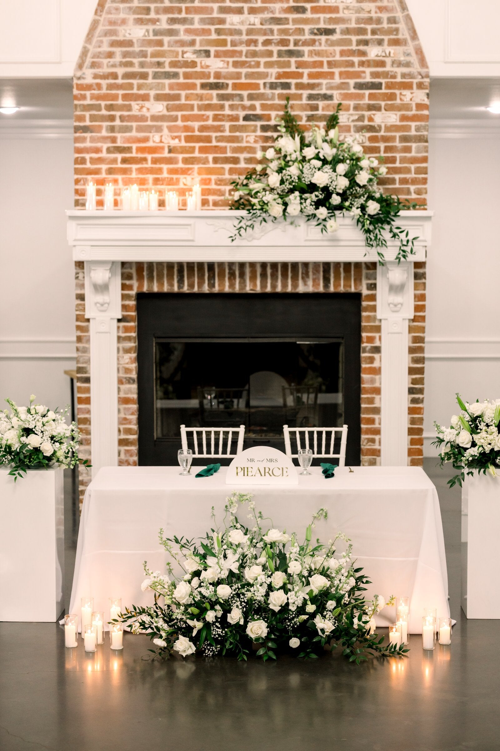Sweetheart table at Parker Manor set against a brick fireplace, with white florals on the mantle and a candlelit ground arrangement at its base by B.blumen