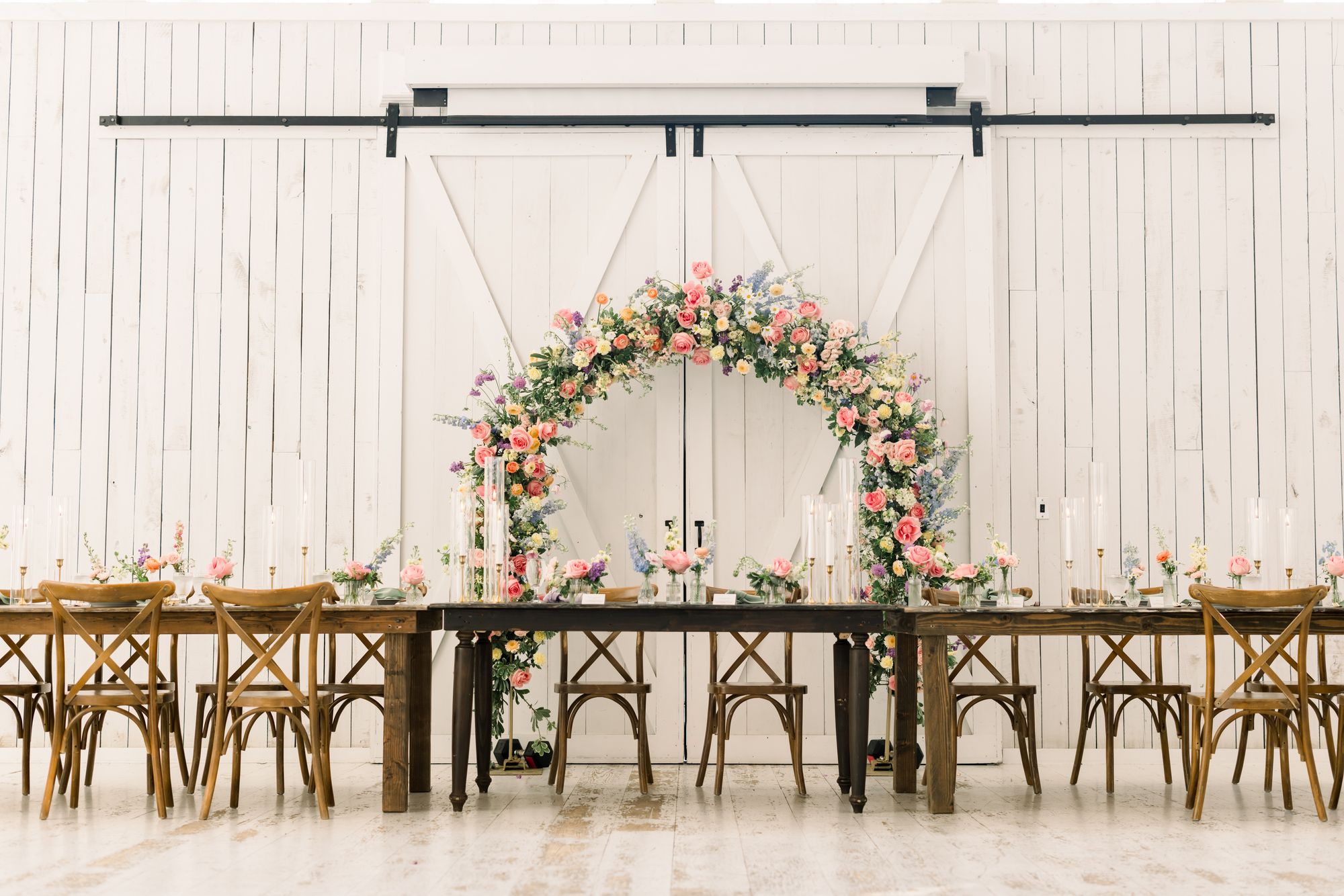 Outdoor ceremony arch at The White Sparrow set against the barn&rsquo;s white sliding doors, pink and green florals over the paved aisle with cross-back chairs, by B.blumen