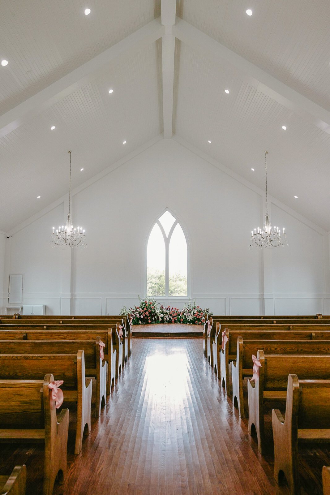 Symmetric view down the wood-pew aisle of The White Sparrow chapel to the arched window, blush aisle florals, by B.blumen