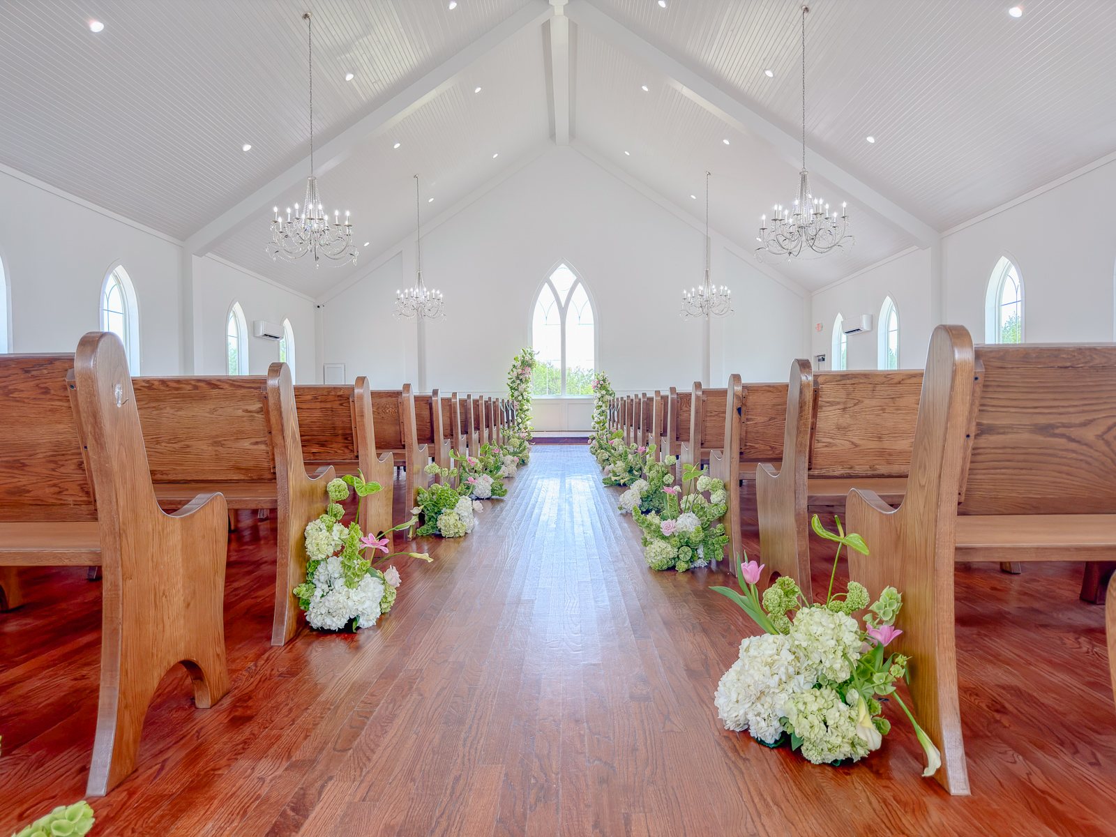 Chapel at The White Sparrow, vaulted white ceiling and chandeliers, wood pews, white and green aisle florals leading to the arched window, by B.blumen