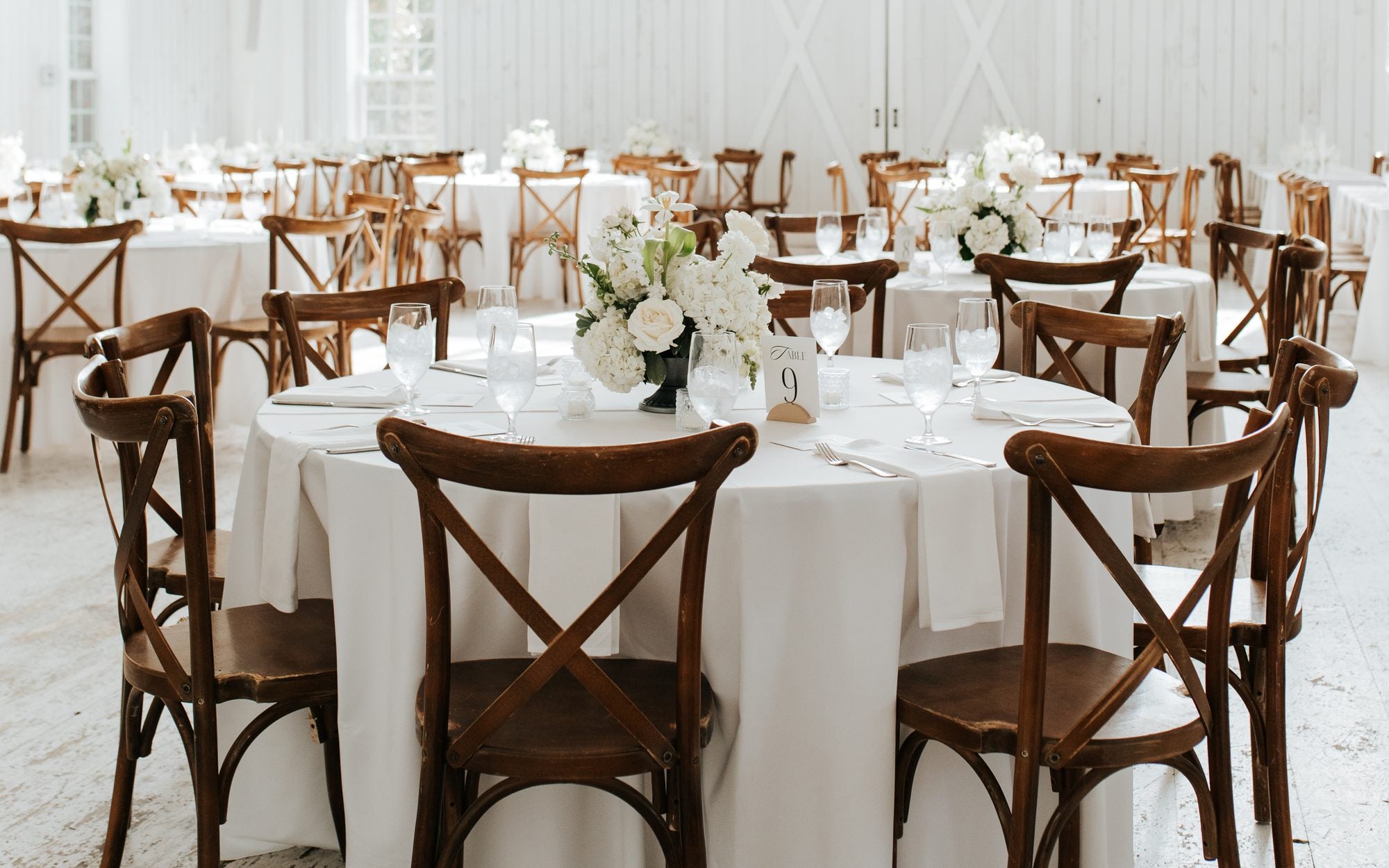 Reception inside The White Sparrow, a round table with low florals and a tall gabled window behind, whitewashed barn interior, by B.blumen