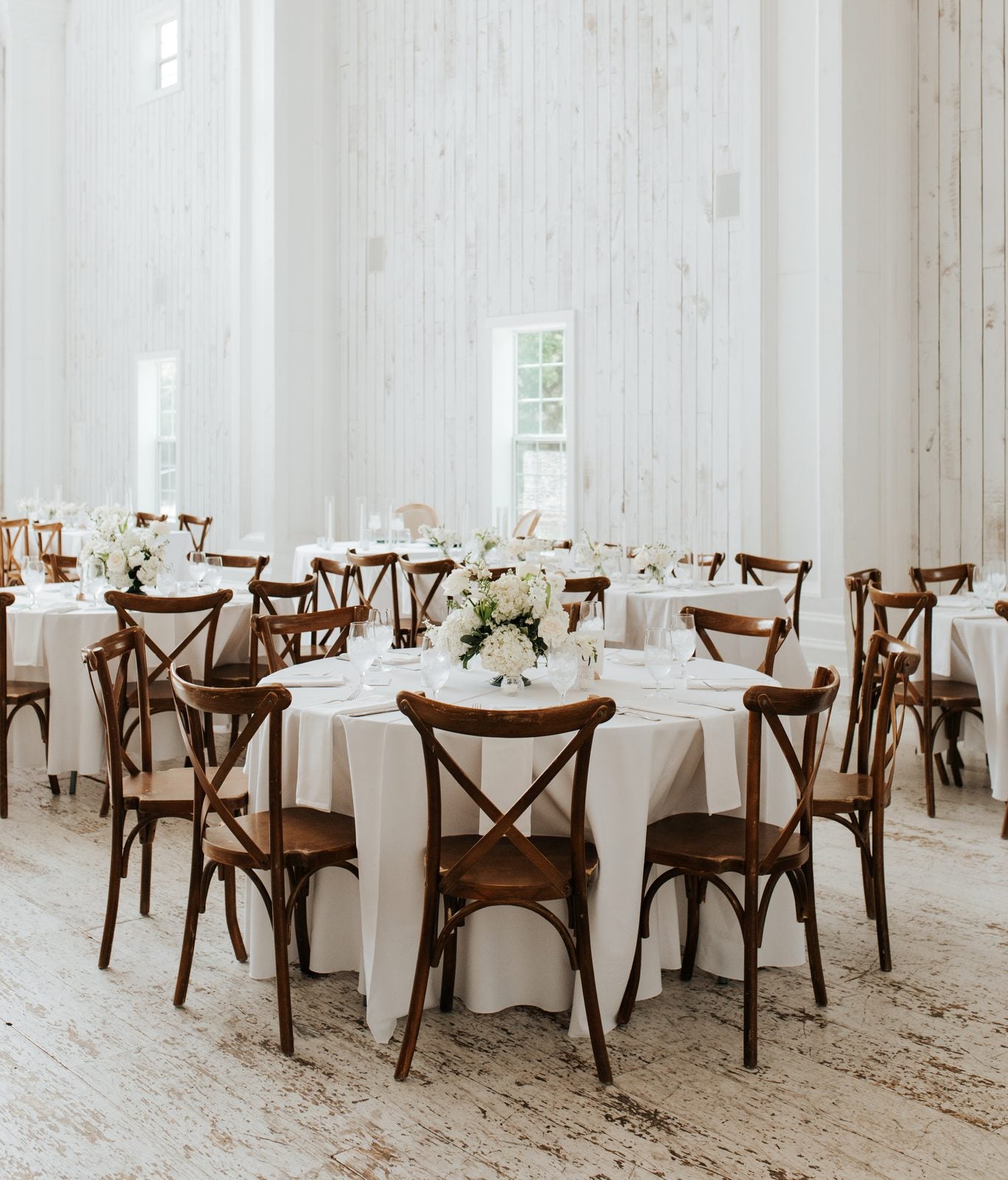 Round table at The White Sparrow with low white floral centerpiece, cross-back chairs, and whitewashed shiplap walls behind, by B.blumen