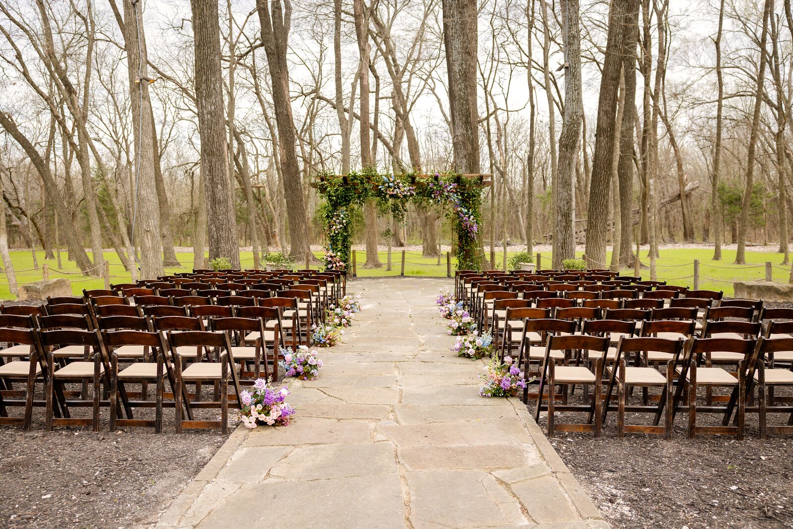 Outdoor ceremony space at The Springs Waxahachie with rows of wooden chairs facing an ivy-covered pergola altar by B.blumen