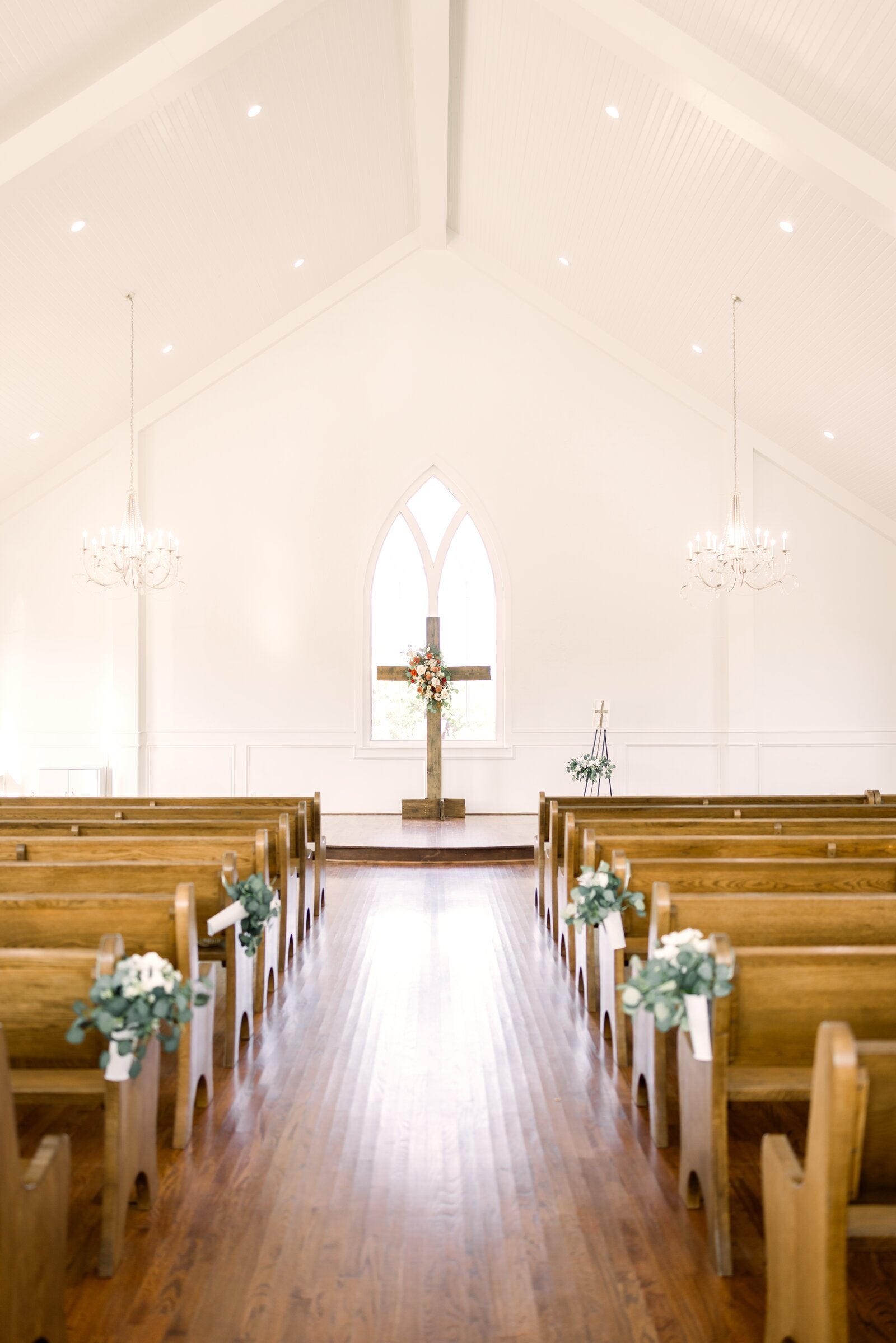 Chapel altar at The Springs Weatherford with wood cross and floral arrangement framed by the gothic arched window, eucalyptus aisle markers down the pews by B.blumen