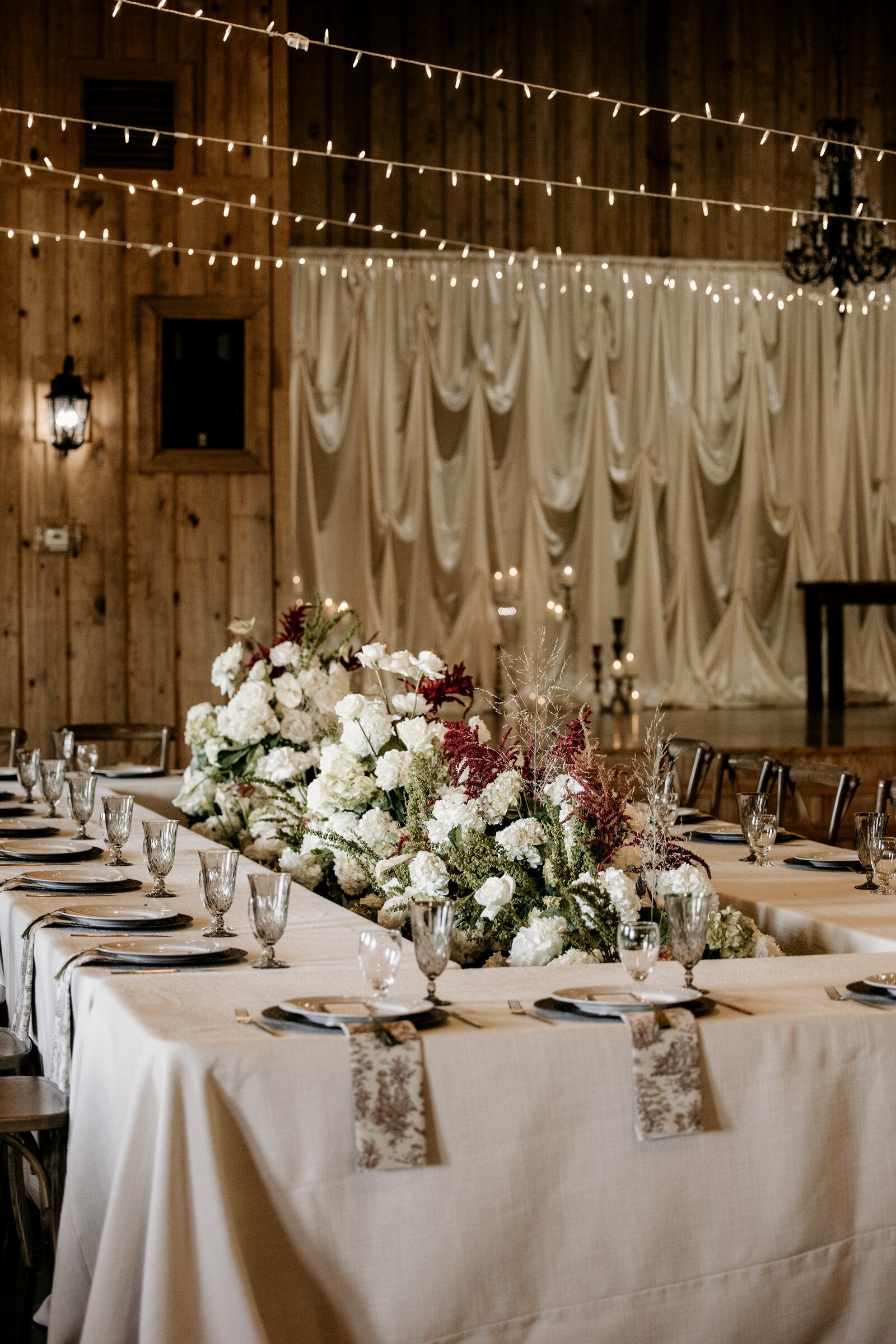 Long reception table at Westwood Hall with white and burgundy centerpiece, draped backdrop, and string lights overhead by B.blumen